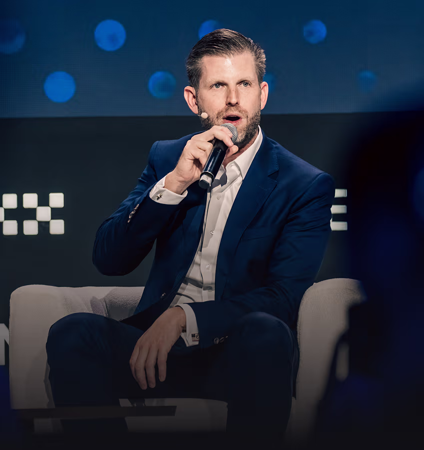 Man in a blue suit speaking into a microphone while sitting on a stage chair.